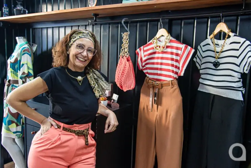 Mulher branca em pé ao lado de looks na sua loja de roupas. Ela é dona de uma loja pertinho de casa.