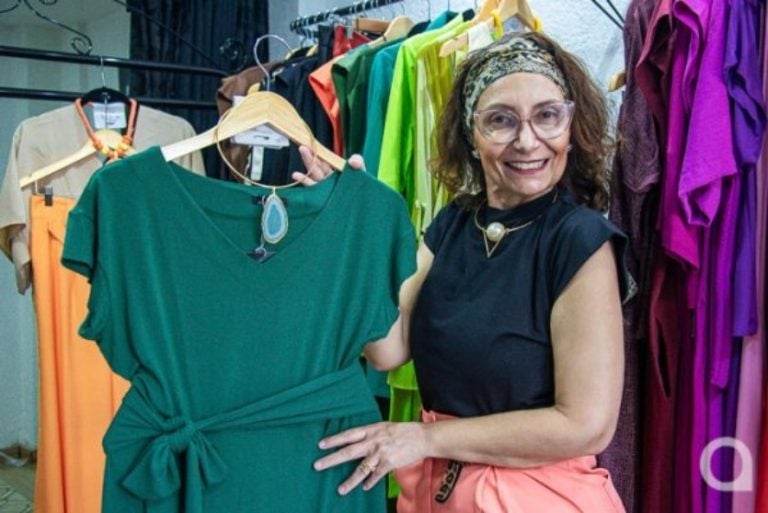 Mulher branca em pé segurando um vestido na cor verde. Ela é dona de uma loja pertinho de casa.