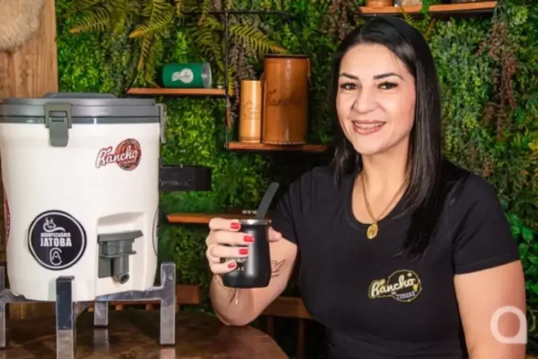 Foto de uma mulher branca de cabelos pretos ao lado de uma jarra e segurando uma cuia de tereré em sua loja especializada em erva mate.