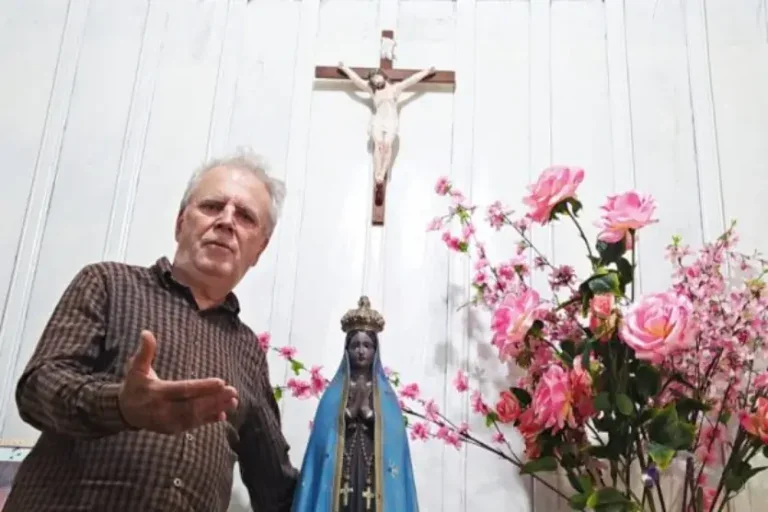 Homem granco em pé ao lado de uma imagem da santa católica Nossa Senhora Aparecida dentro de uma capelinha rural