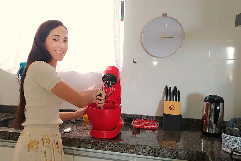 Mulher branca de cabelos pretos sorrindo parada em frente a uma batedeira vermelha mexendo na vasilha com uma espátula em uma cozinha