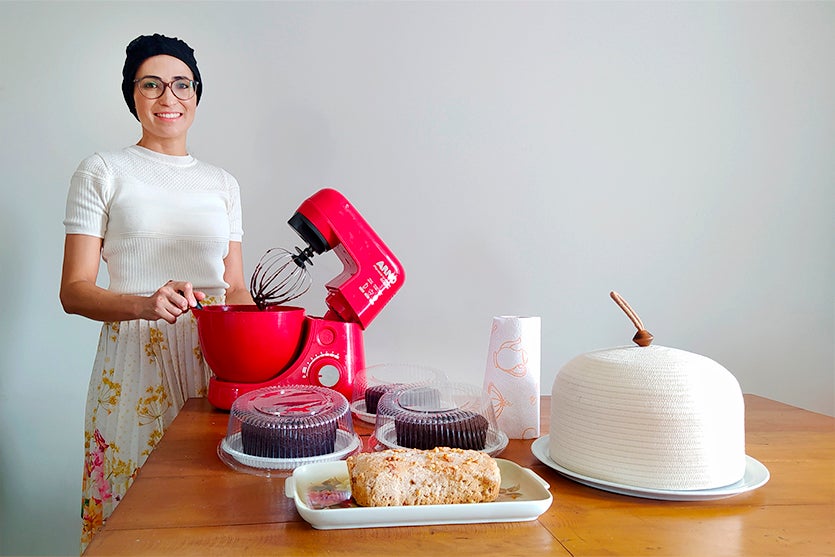 Mulher branca com lenço na cabeça ao lado de batedeira vermelha. Na mesa também tem bolos sem glúten e sem lactose para intolerantes
