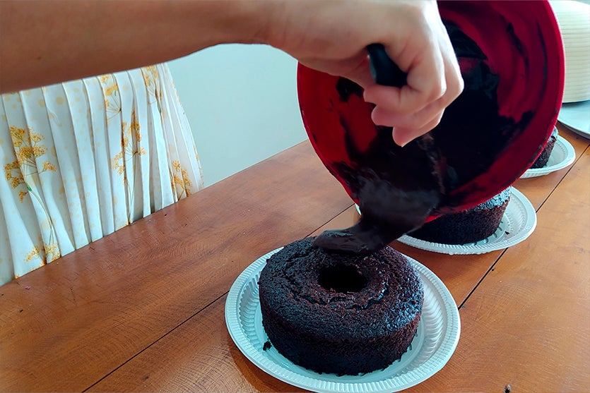 Mulher cobrindo um bolo de chocolate com cobertura de chocolate que está derramando de um vasilha vermelha e usando uma espátula de silicone