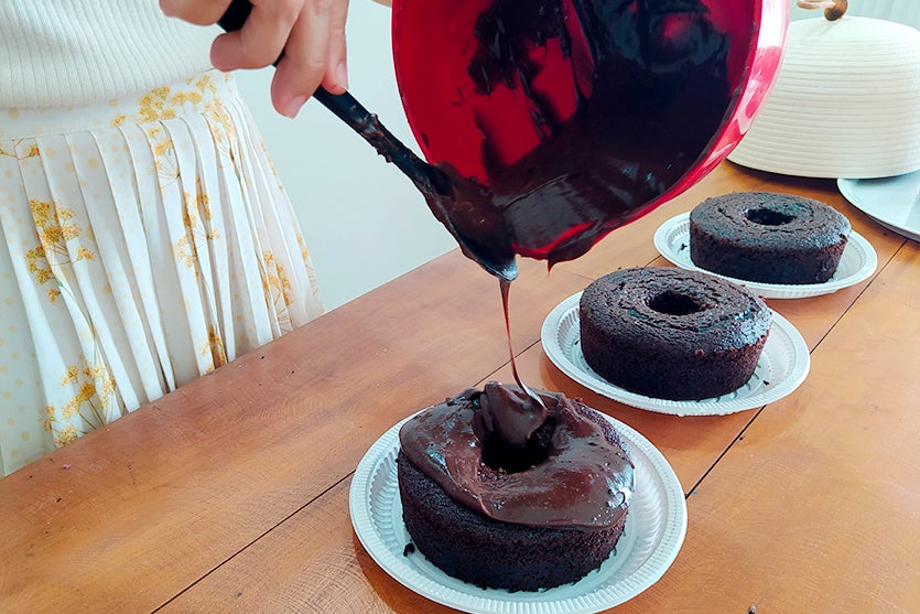 Mulher cobrindo um bolo de chocolate com cobertura de chocolate que está derramando de um vasilha vermelha e usando uma espátula de silicone