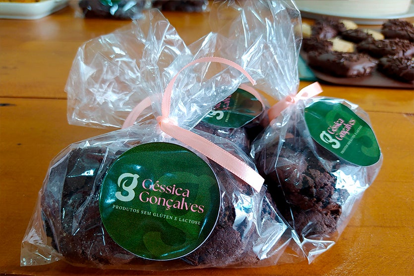 Embalagem transparente com cookies sem glúten e sem lactose para intolerantes dentro amarrados com um laço rosa e etiqueta verde com nome da boleira sobre uma mesa marrom.