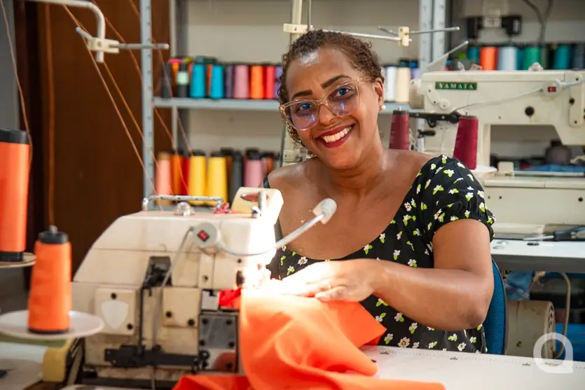 Mulher negra usando óculos de grau e sorrindo sentada em frente a uma máquina de costura costurando uma roupa cor de laranja
