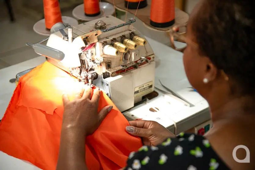 Mulher negra usando óculos de grau sentada em frente a uma máquina de costura costurando uma roupa cor de laranja