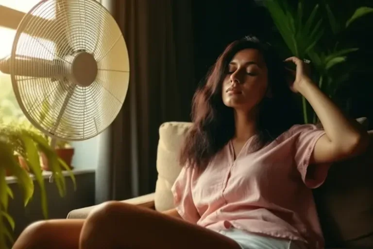 Mulher sentada em frente a um ventilador com os olhos fechados e uma mão na cabeça. Foto usada para ilustrar matéria sobre como deixar a casa fresca sem ar-condicionado