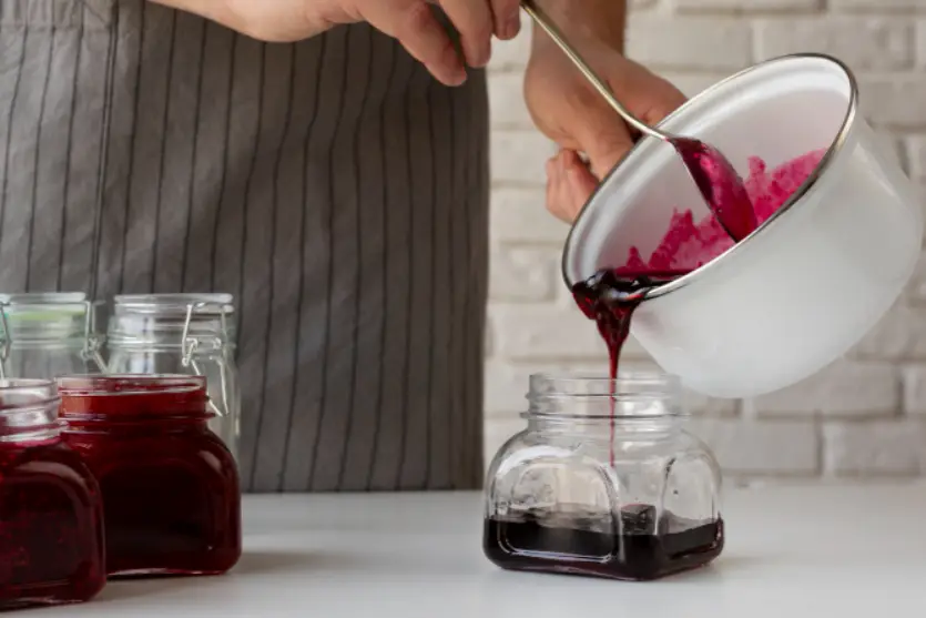Mãos de uma mulher branca colocando um geléia de frutas vermelhas em um pote transparente