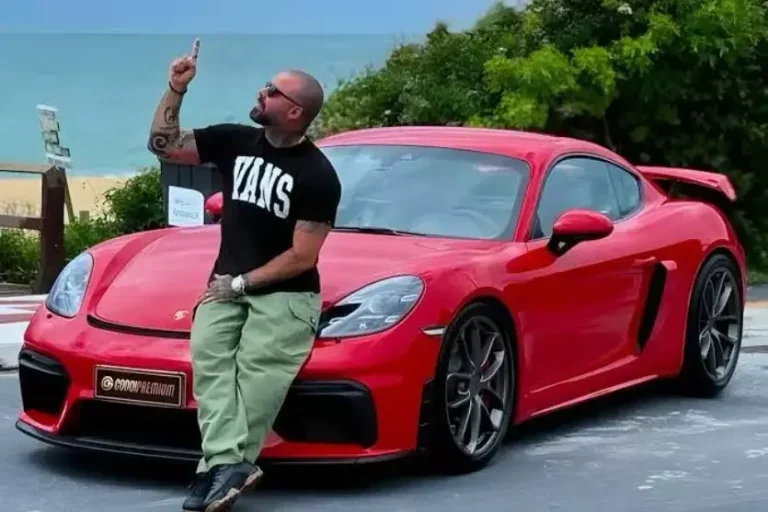 Influenciador Ricardo Godoi é um homem branco com camisa preta sentado sobre um carro vermelho de luxo.