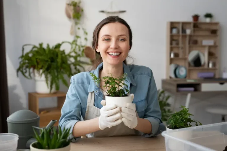 Mulher branca sorrindo e segurando um vaso de plantas branco com luvas brancas dentro de uma casa. Foto para ilustrar a matéria sobre jardinagem para iniciantes.