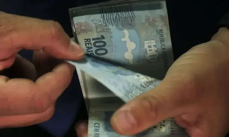 Foto de uma mão de uma pessoa branca contando notas de cem reais. Imagem usada para ilustrar a matéria sobre o pagamento do abono salarial Pis/Pasep 2025.