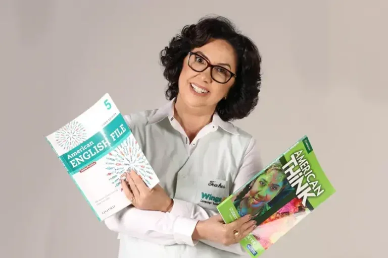 Foto da professora que da cicas de inglês gratuitas em grupo de whatsapp. Ela é uma mulher branca de cabelos pretos e que usa óculos. Ela está em pé, sorrindo e segurando livros de inglês.