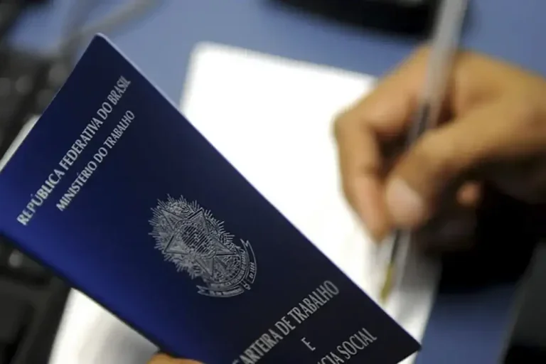 Carteira de trabalho aberta sendo segurada por uma mão de homem que está escrevendo com a outro mão em um papel branco. Foto para ilustrar matéria sobre reforma trabalhista.