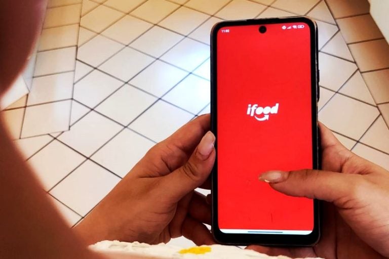 Valor mínimo para pedidos do iFood. Na foto, uma mão feminina segurando o celular com a tela do aplicativo.