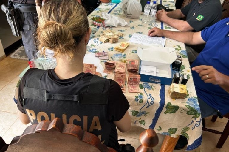 Fraudes no programa Farmácia Popular. Na foto, uma policial checando documentos em uma mesa com dinheiro.