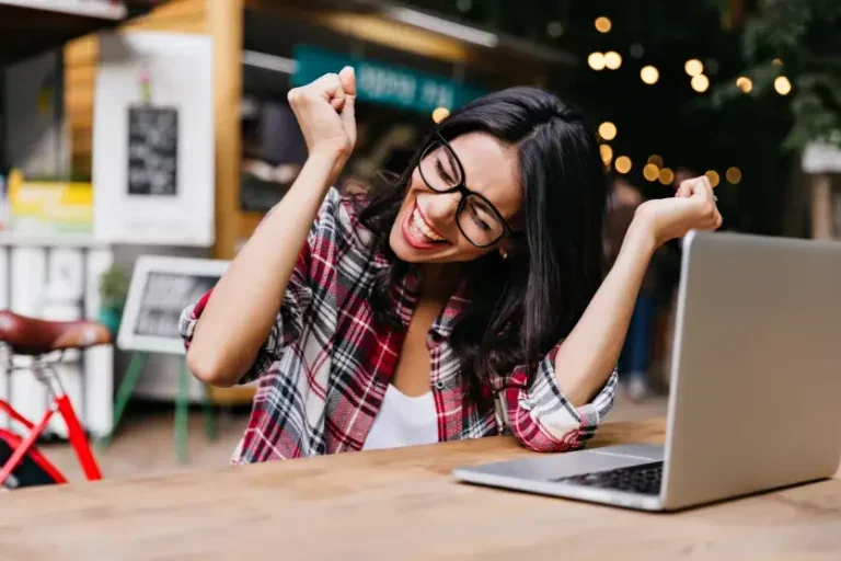 Mulher branca de cabelos pretos usando óculos sentada em frente a um notebook comemorando. Foto para ilustrar uma matéria sobre o concurso da cesgranrio.