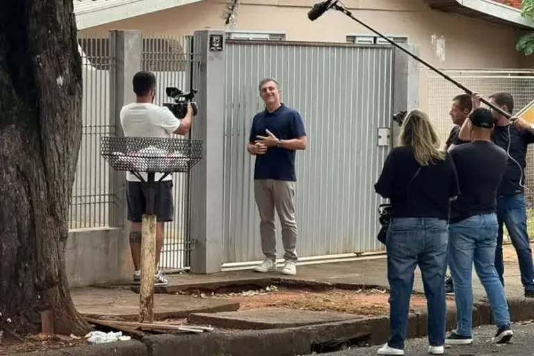 Luciano Huck em frente a uma casa com portão cinza com uma equipe de filmagem.