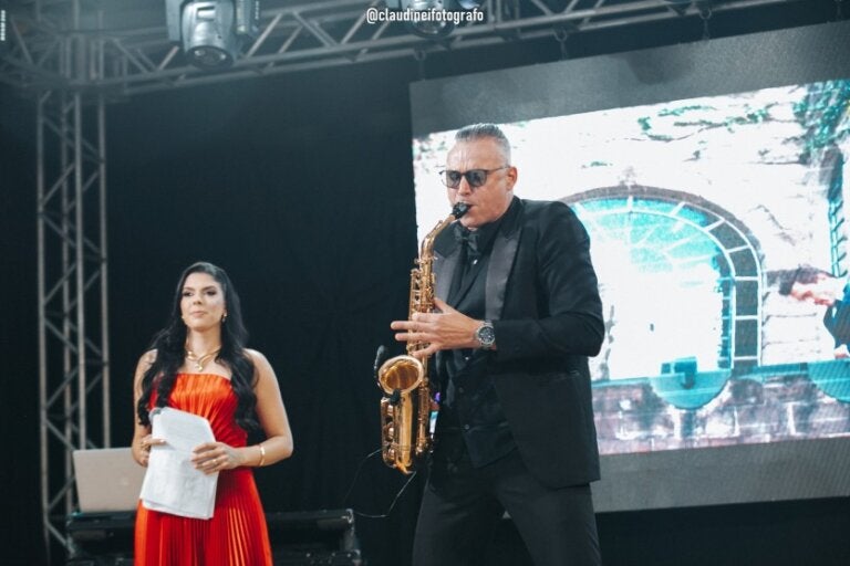 Saxofonista que irá tocar na Black Prime Wedding durante apresentação em palco ao lado de uma apresentadora.