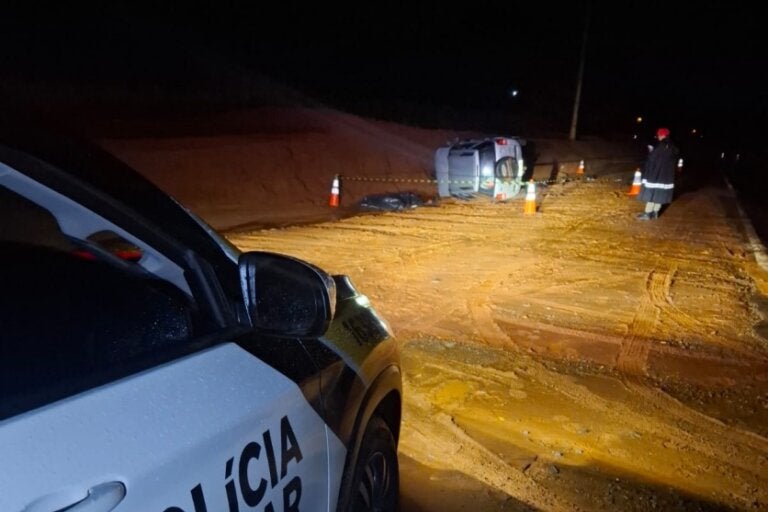 Foto de uma viatura policial no local do acidente em que um motorista morreu após colidir contra um barranco na PR-464, em Inajá. Corpo da vítima está coberto em uma lona preta.