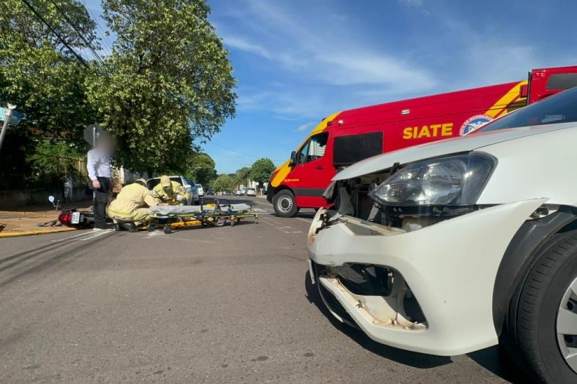 Local do acidente entre uma moto e um carro que resultou em um motociclista com a perna fraturada, em Umuarama. Há o veículo envolvido na batida e ao fundo a vítima recebendo atendimento do Siate.