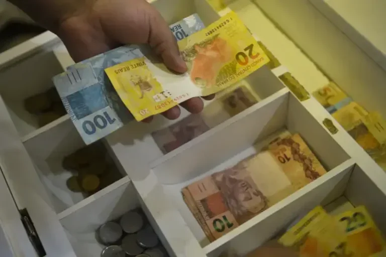 Foto de uma mão com notas de dinheiro em frente a uma caixa registradora.