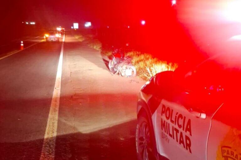 Foto de uma viatura da PRE próxima ao local do acidente envolvendo um carro e uma motocicleta na PR-323, entre Umuarama e Perobal. A moto está caída na rodovia.