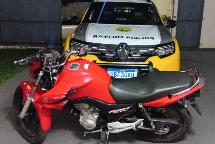 Moto apreendida próxima a uma viatura da PM para ilustrar a matéria sobre o motociclista que fugiu de uma abordagem em Cruzeiro do Oeste.
