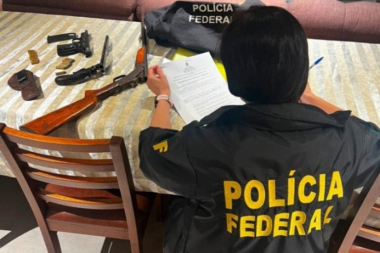 Policial federal durante cumprimento das Operação Sicarius I e II. Ela está analisando um documento próxima a armas de fogo.