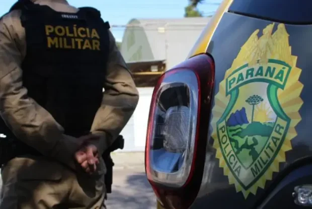 Policial militar usando colete próximo a uma viatura da PM.